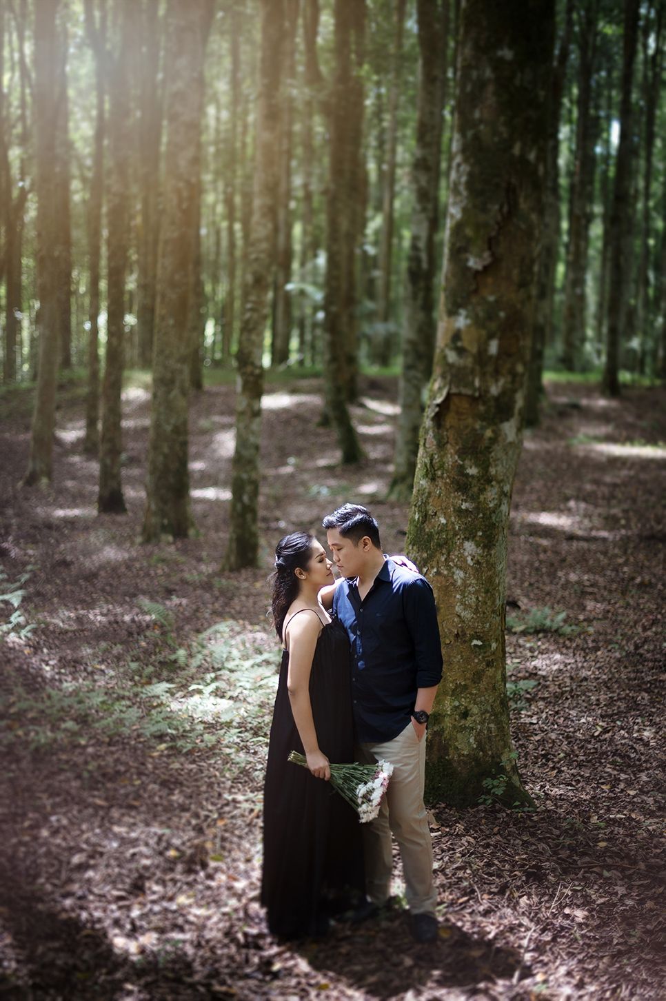 Romantic couple sharing an intimate moment among tall trees in a sunlit forest during a photo shoot.