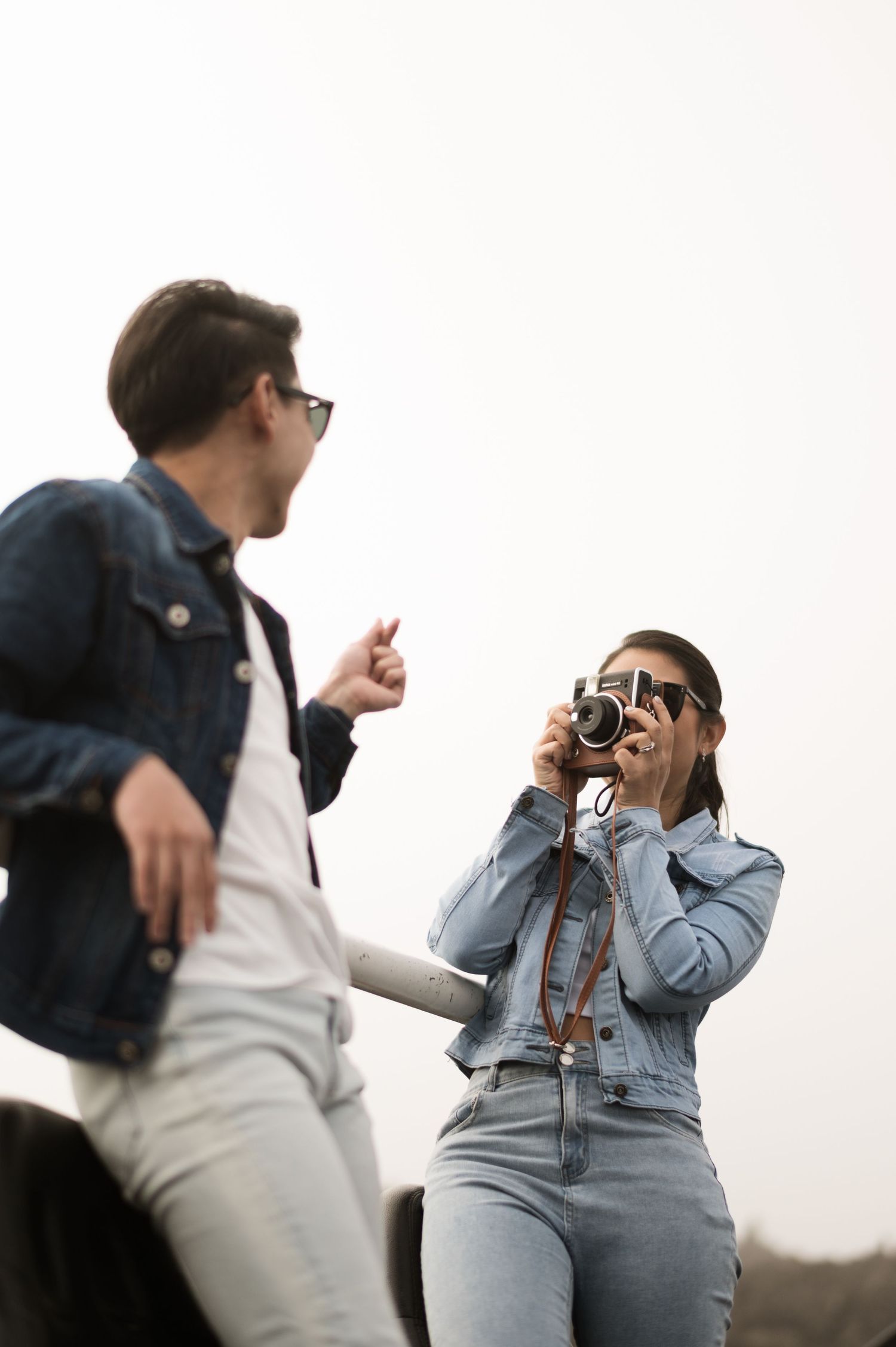 Woman in denim jacket photographs man outdoors using vintage camera, both wearing sunglasses on overcast day.