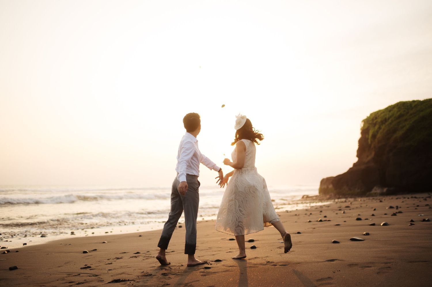 Couple dancing on a sandy beach at sunset, holding hands near ocean waves and rocky cliffs.