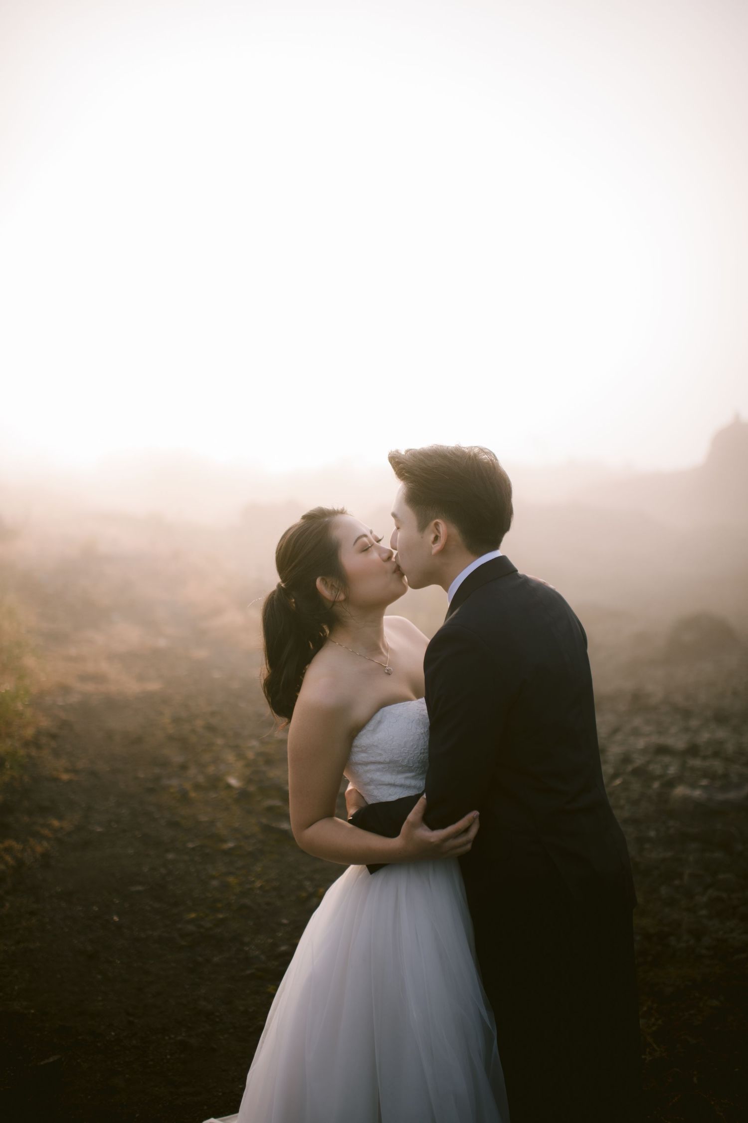 A bride and groom share a romantic kiss in a misty outdoor setting, the bride in a white gown and groom in a dark suit.
