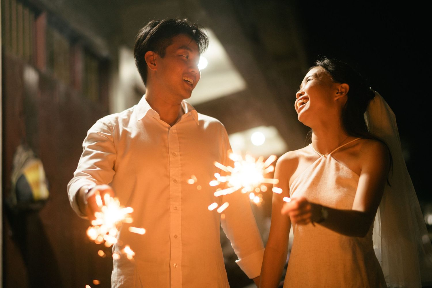 A joyful Asian couple in wedding attire holds sparklers together, smiling at each other in a romantic nighttime setting.