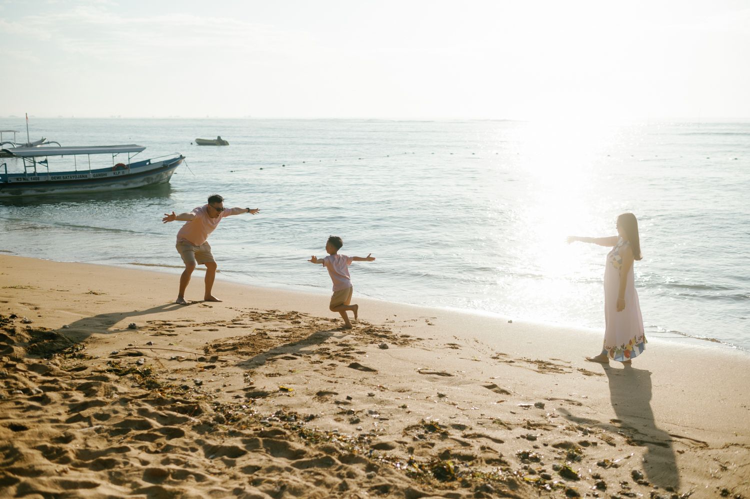 Family playing on a sunny beach with boats in the background, child running toward parent with arms outstretched.