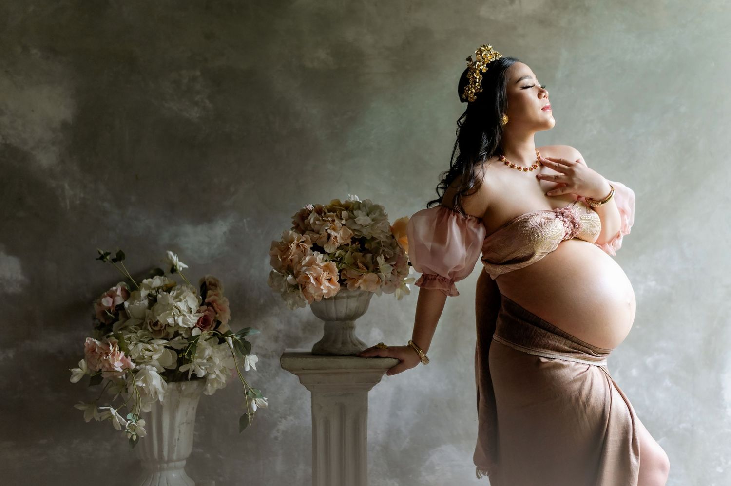Elegant maternity photo of a pregnant woman in a pink gown wearing a crown, posed beside floral arrangements in a studio.