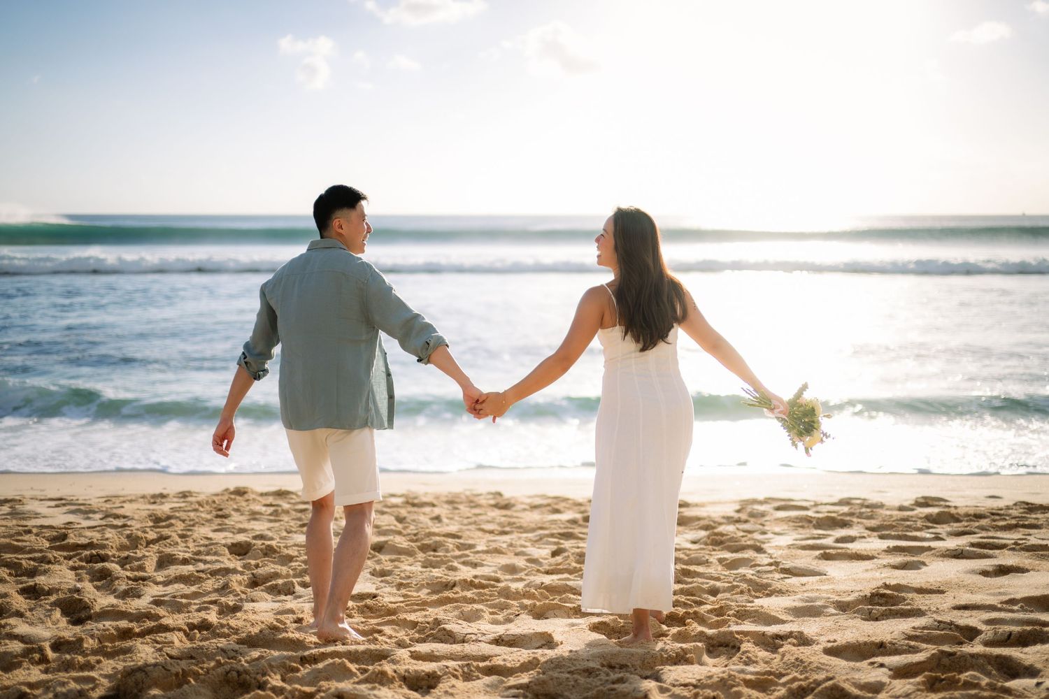 Couple holding hands on a sandy beach at sunset, woman in white dress holding flowers, man in casual attire.