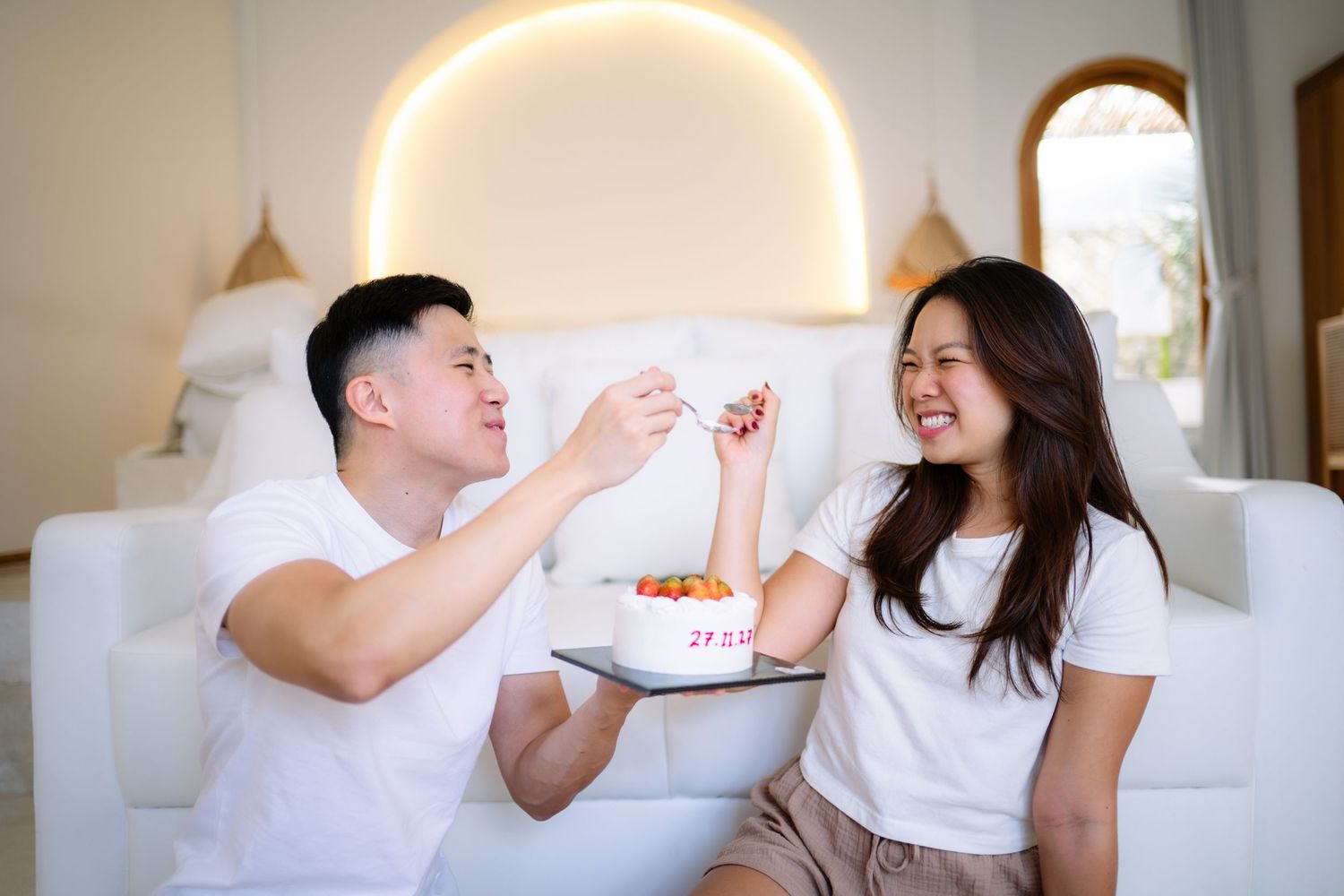 Couple sharing cake on white sofa, man feeding woman who smiles, cozy home setting with warm lighting.