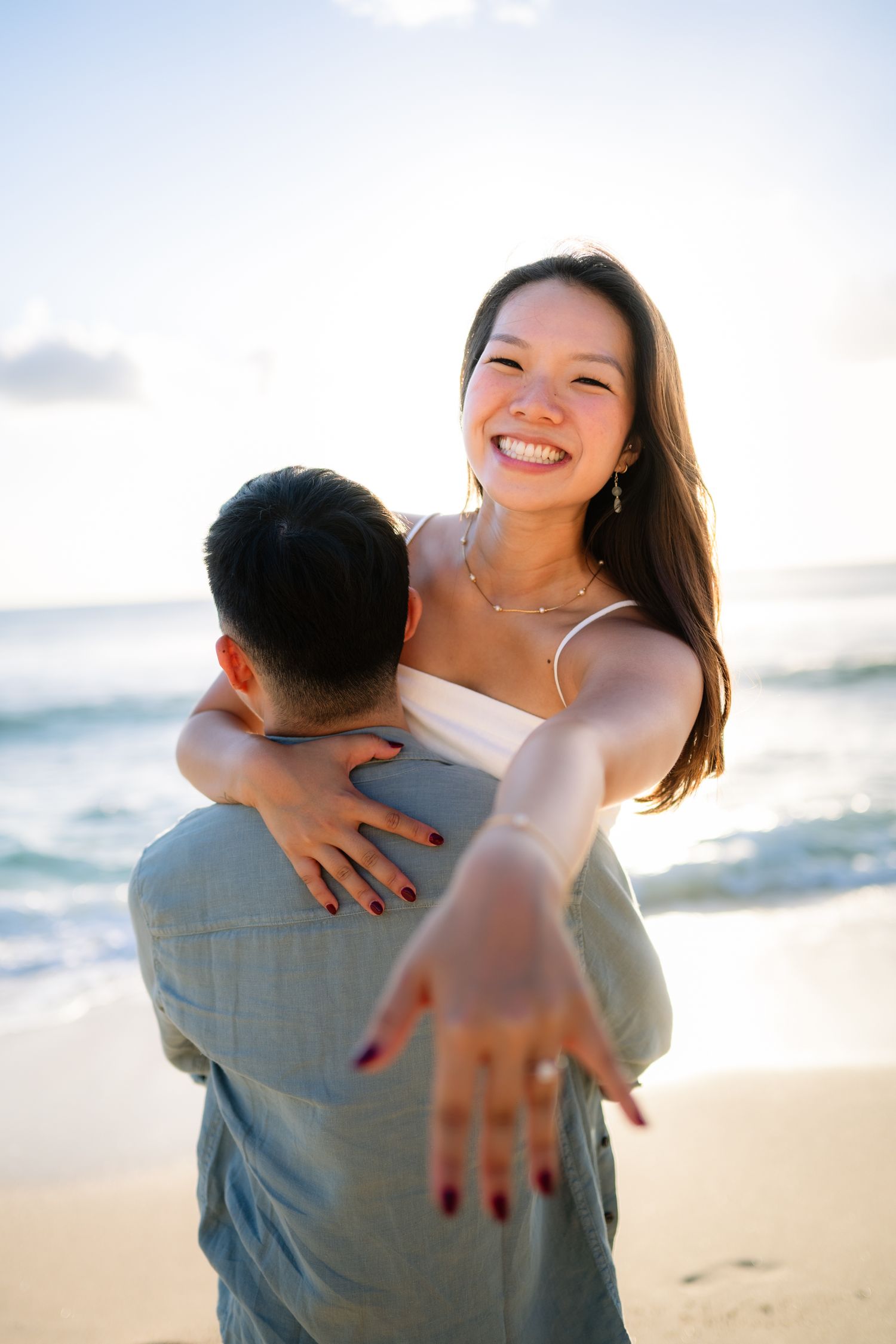 Smiling woman showing engagement ring while being lifted by partner on a sunny beach.