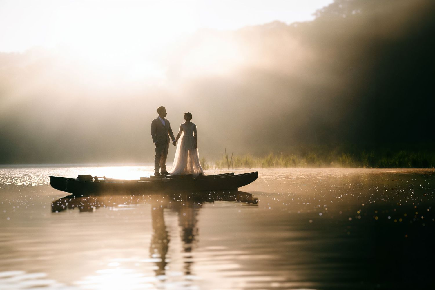 Couple holding hands standing on a boat at sunset on a misty lake with golden light reflecting on the water.