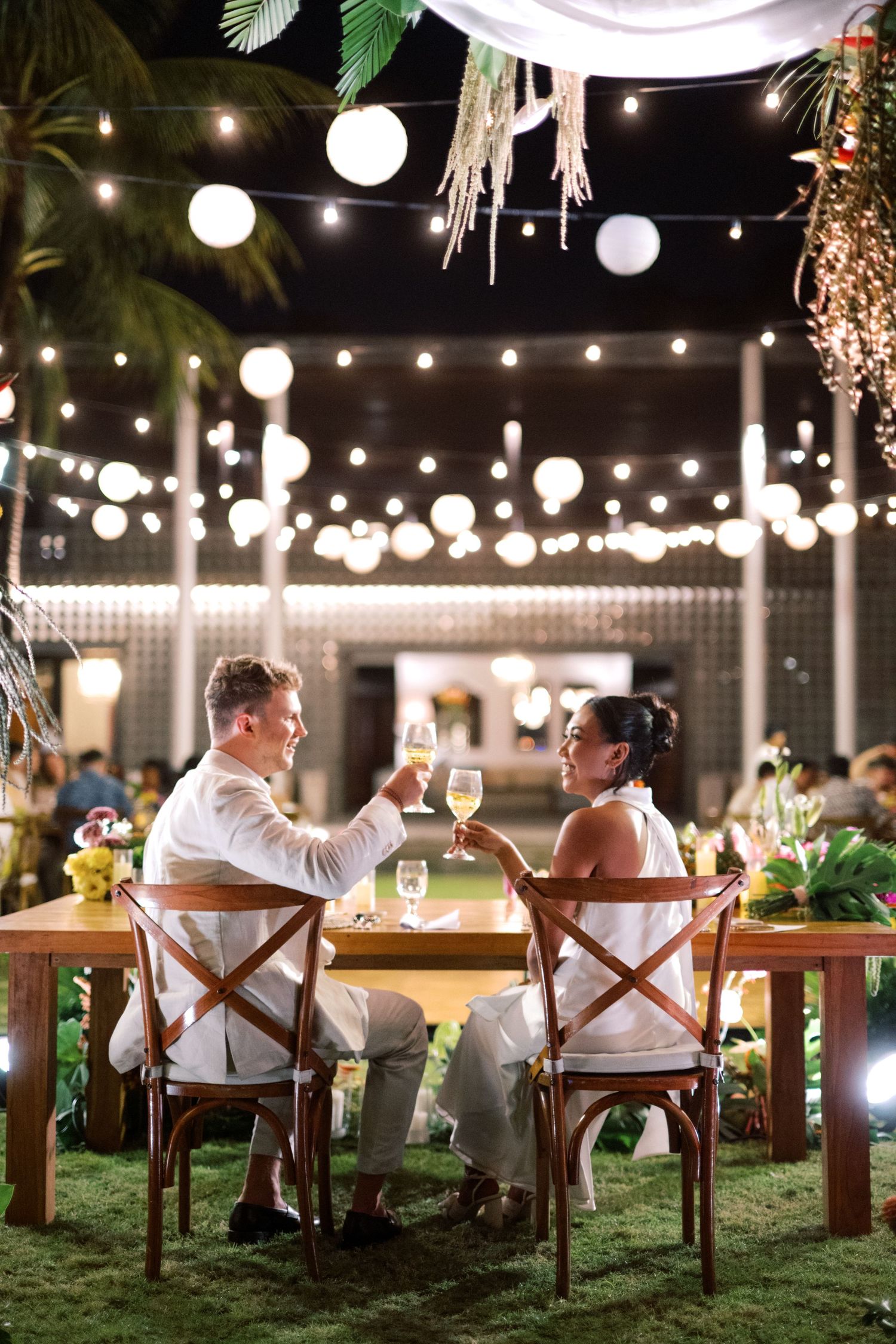 Couple in white attire toasting champagne at outdoor wedding reception with string lights and lanterns at night.