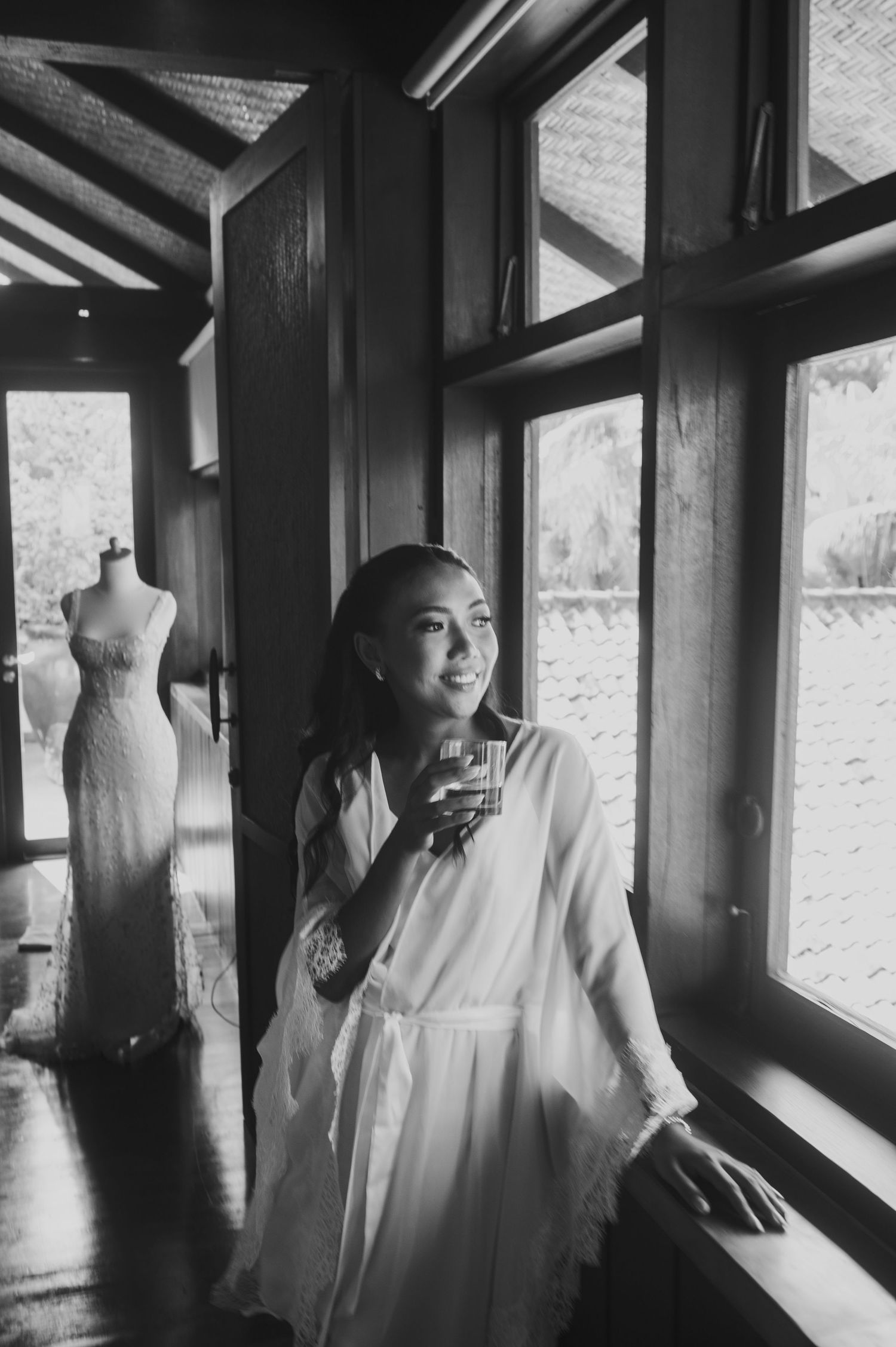 Black and white photo of a smiling bride in a robe holding a drink by a window, with a wedding dress on a mannequin behind her.