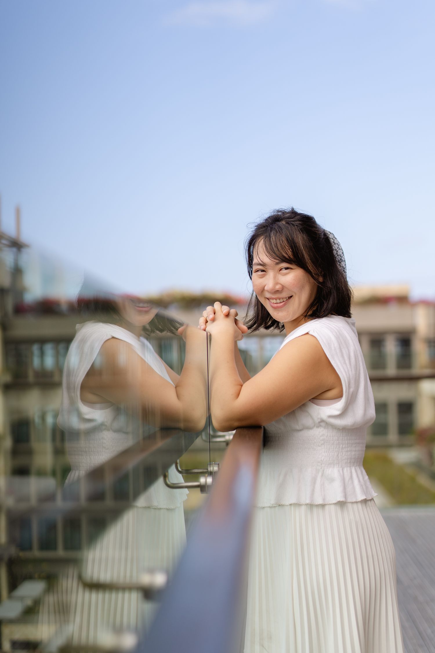 Smiling Asian woman in white dress leaning on glass balcony railing with blue sky and buildings behind her.
