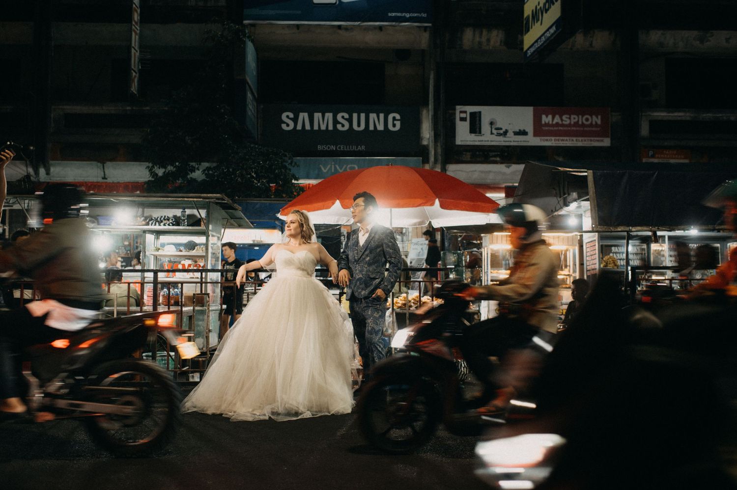 Bride and groom pose on a busy night street in front of a Samsung store, with motorcycles passing by.