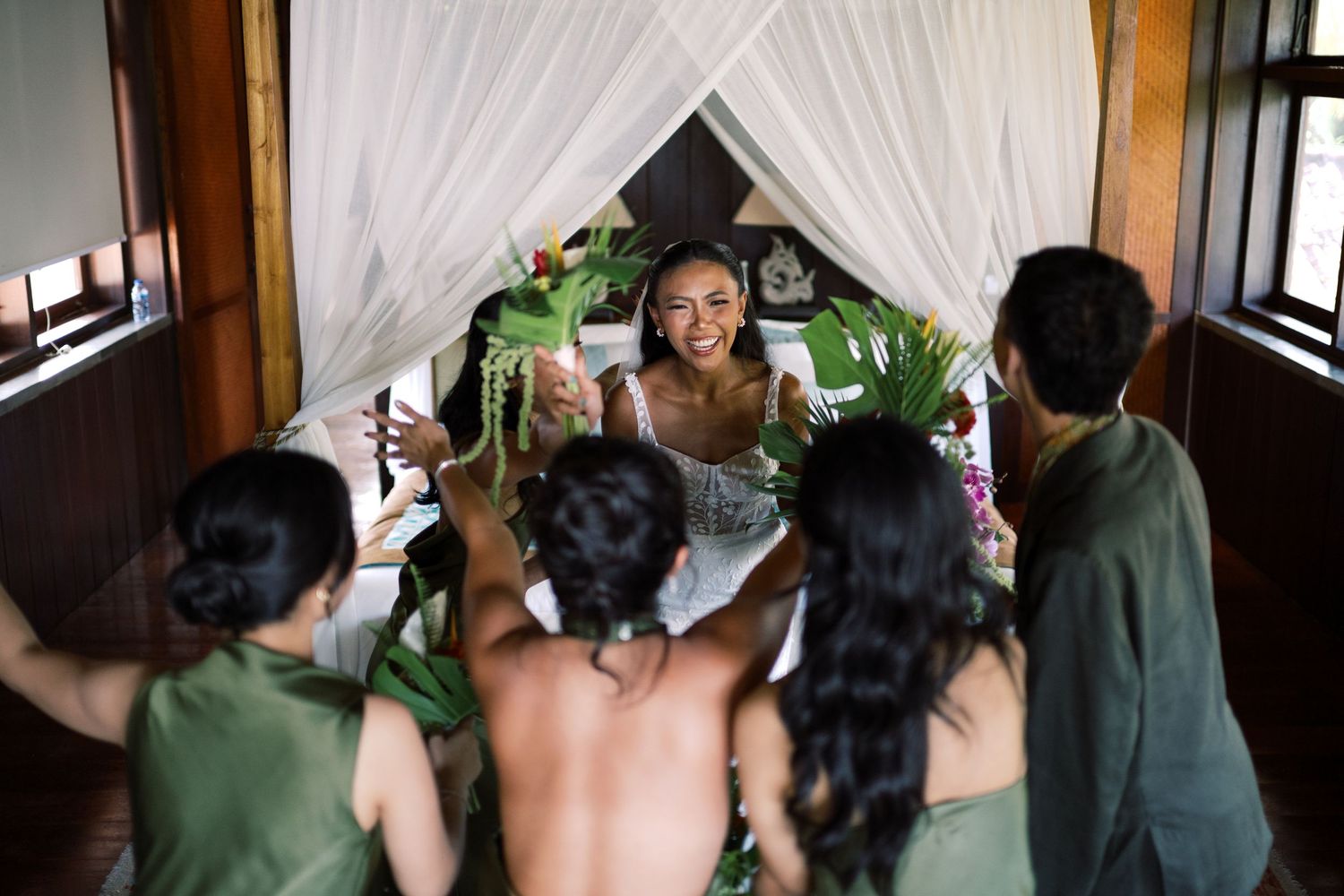 Bride laughs joyfully with bridesmaids holding tropical flowers in decorated wedding suite.