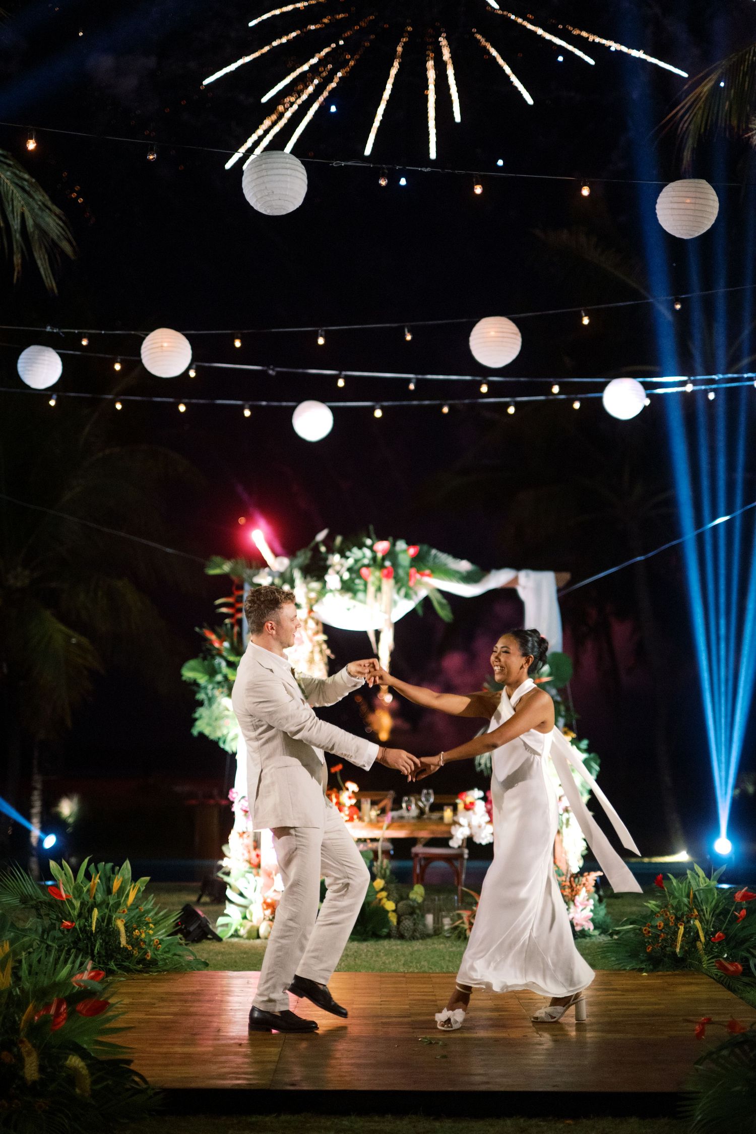 Bride and groom share first dance under fireworks and string lights at tropical outdoor reception.