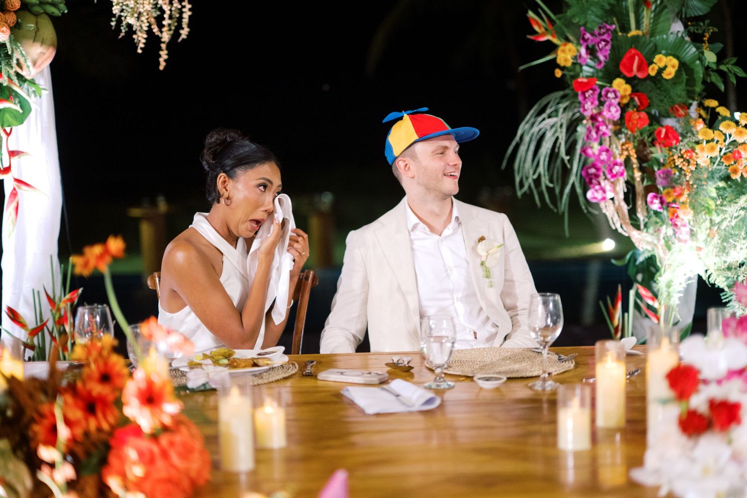 Bride laughs while groom wears a colorful hat at their tropical wedding reception dinner table.