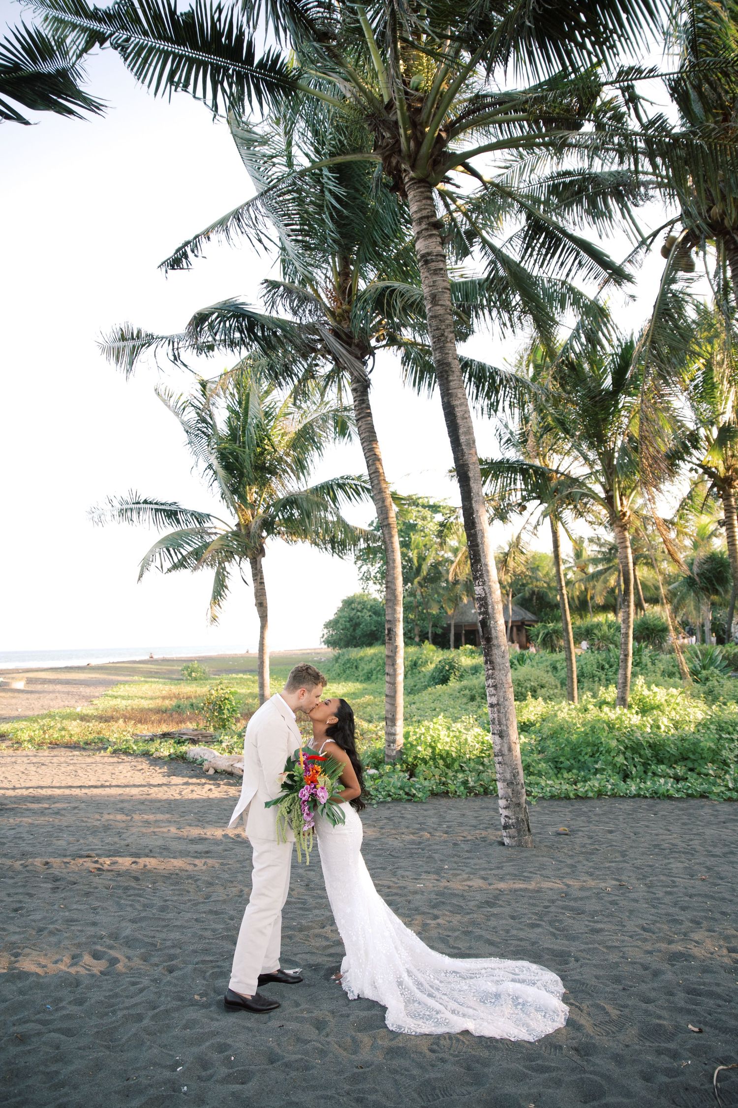 Bride and groom sharing a kiss on black sand beach under swaying palm trees at golden hour.