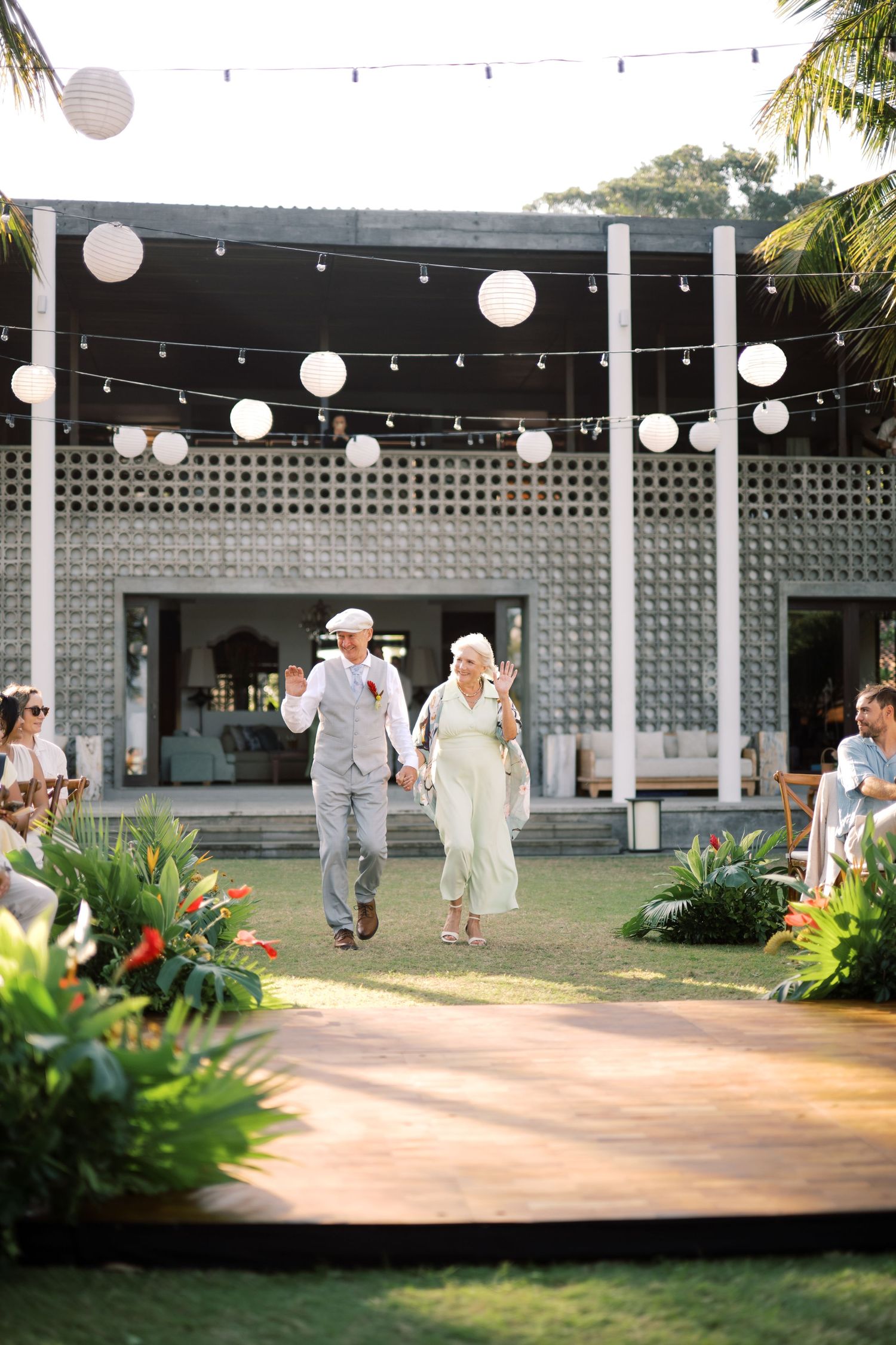 Elderly couple walks down outdoor aisle under string lights at tropical wedding venue.
