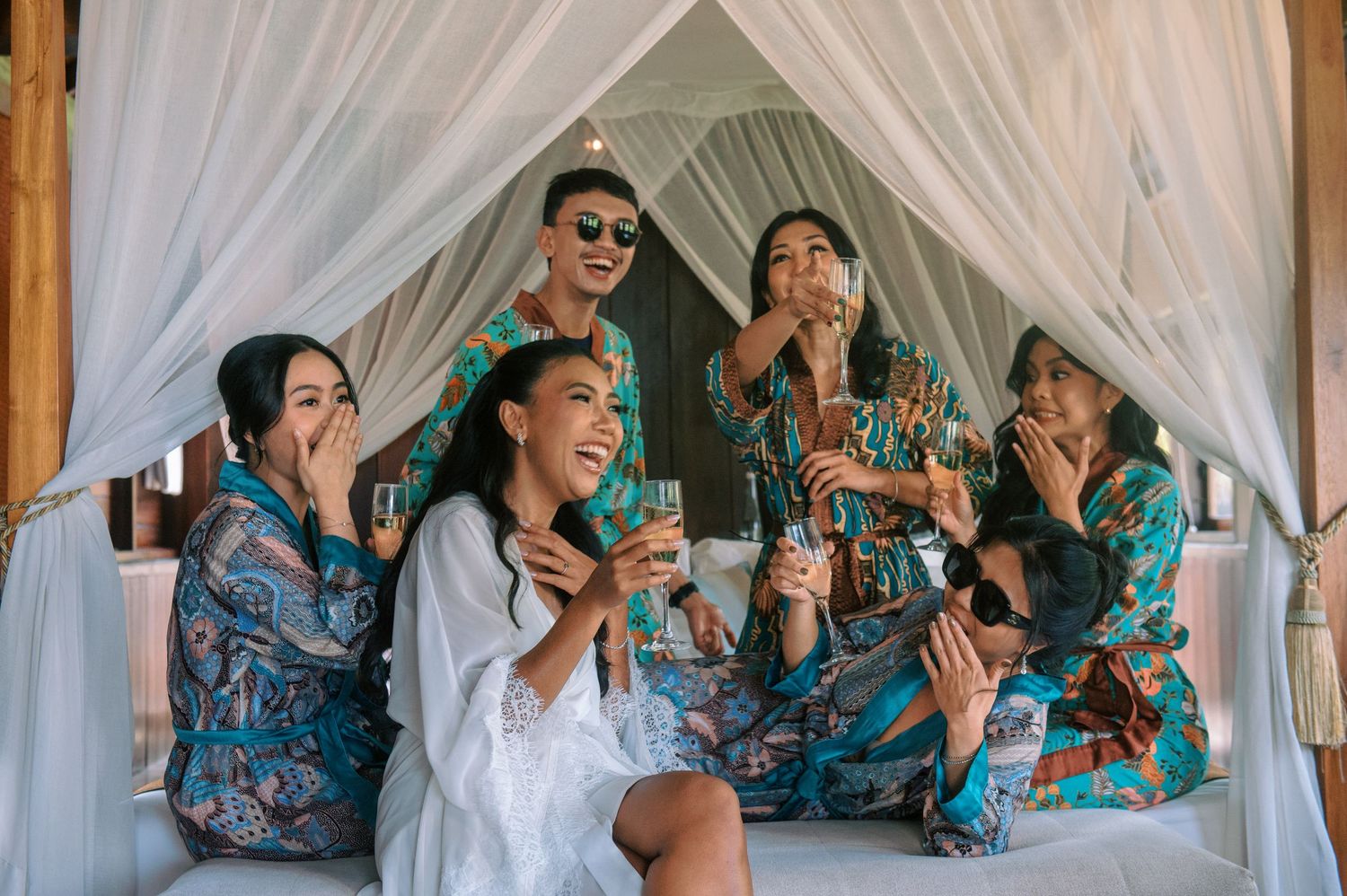 Group of friends in colorful robes celebrating together under a canopy bed, laughing and drinking.