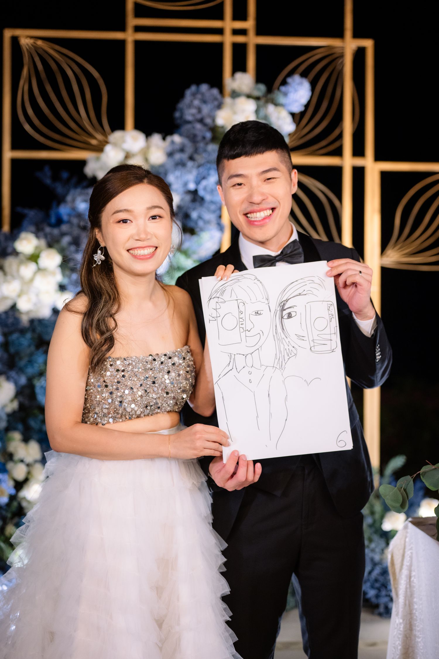 Smiling bride and groom in formal wedding attire hold a caricature sketch of themselves beside blue and white flowers and gold decor.