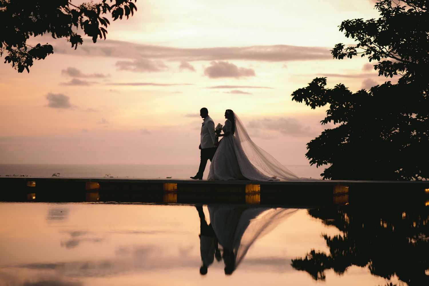 Silhouette of bride and groom at sunset with veil flowing, reflected in calm water at outdoor wedding.