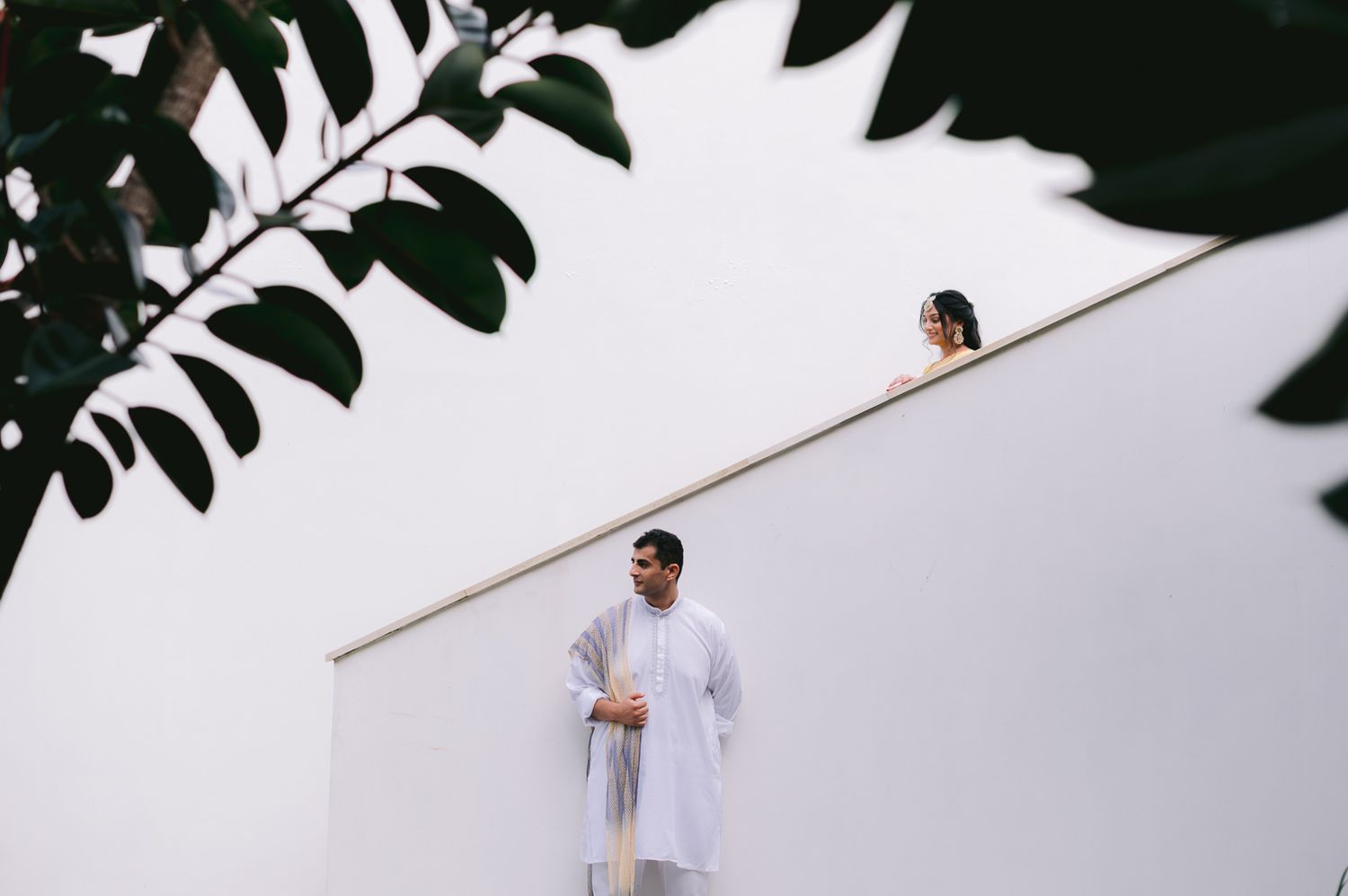 Indian couple in traditional white attire posed on white architectural steps with lush green leaves framing the scene.