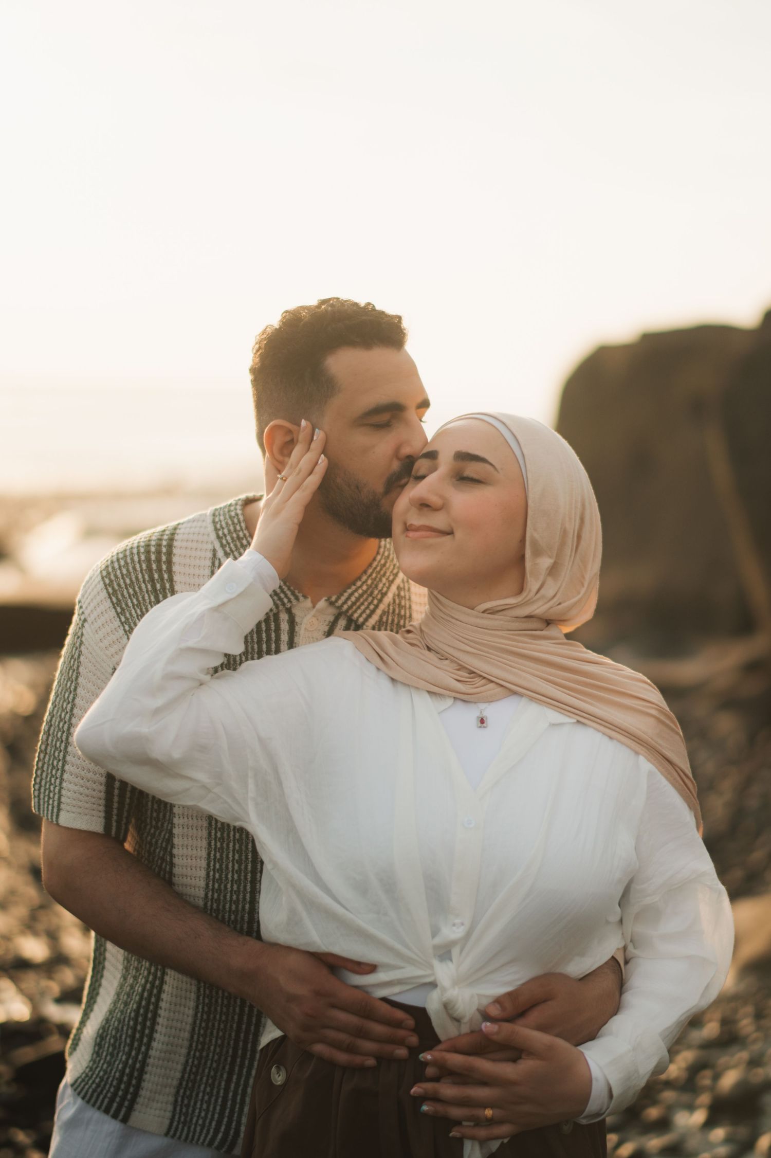 A couple shares a tender moment at sunset, man kissing woman in hijab on cheek near rocky shore.