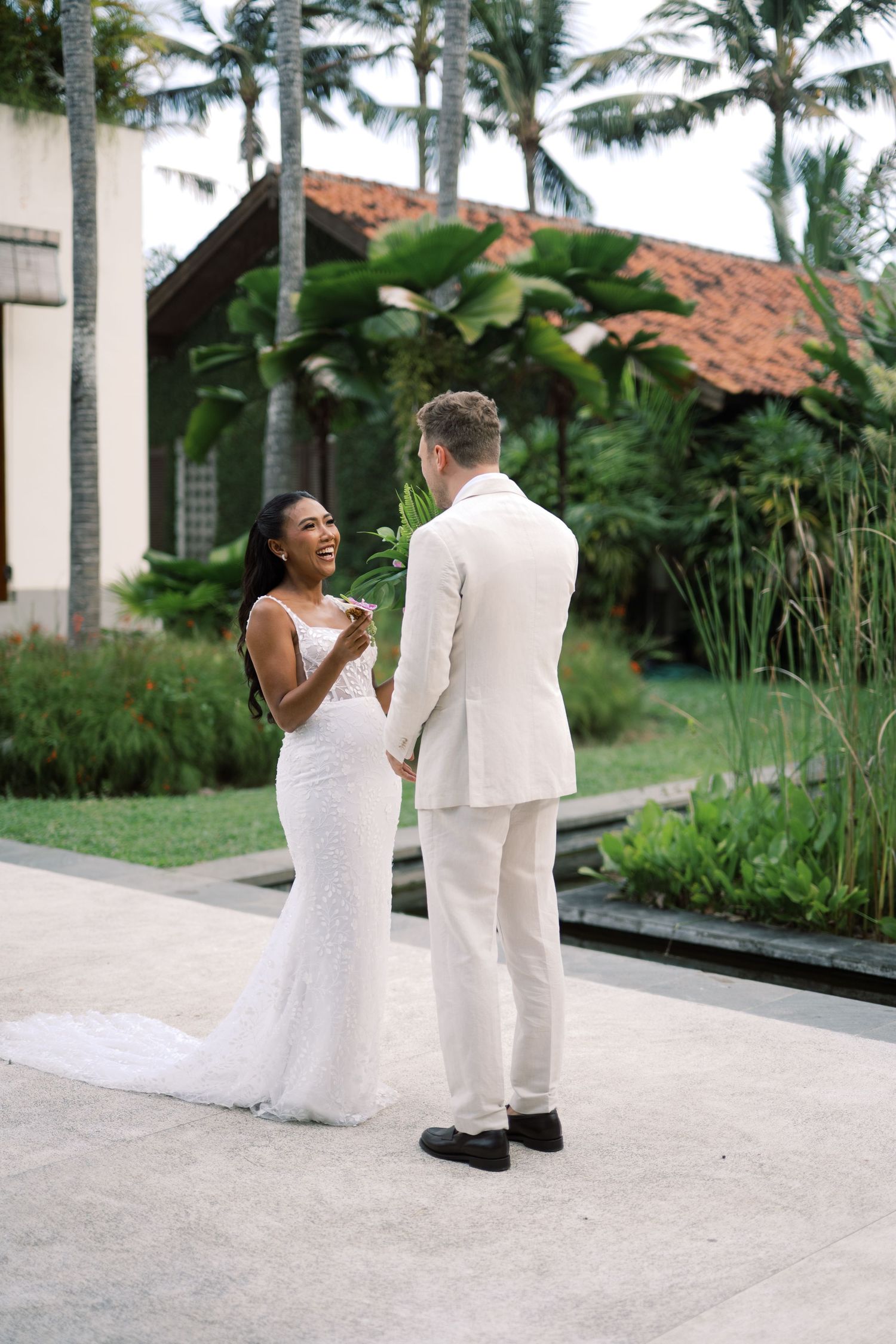 Bride in white lace gown laughs joyfully during first look with groom in cream suit at tropical wedding venue.