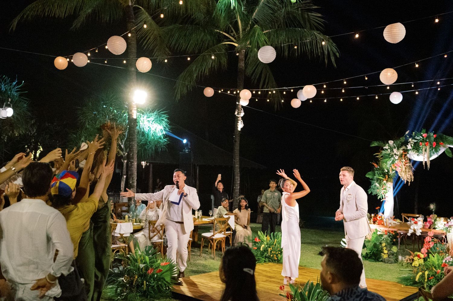 Wedding guests raise glasses in a toast as couple celebrates under string lights at tropical outdoor reception.