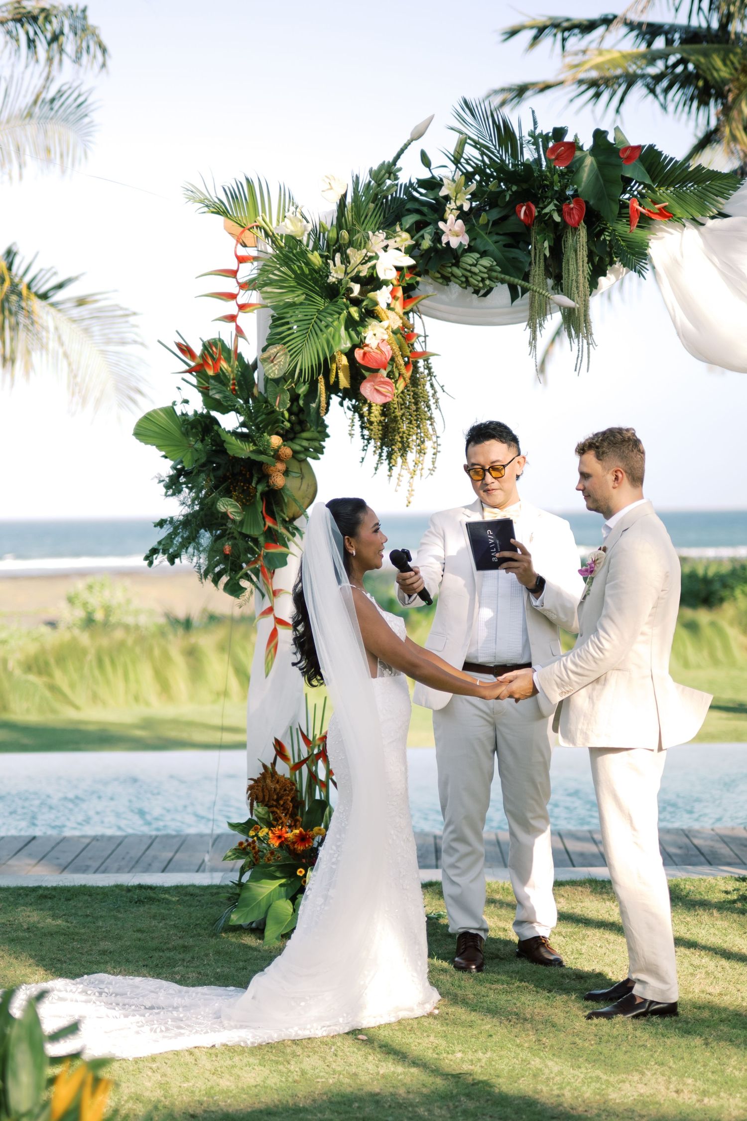Bride and groom exchange vows under tropical floral arch with ocean backdrop and officiant.