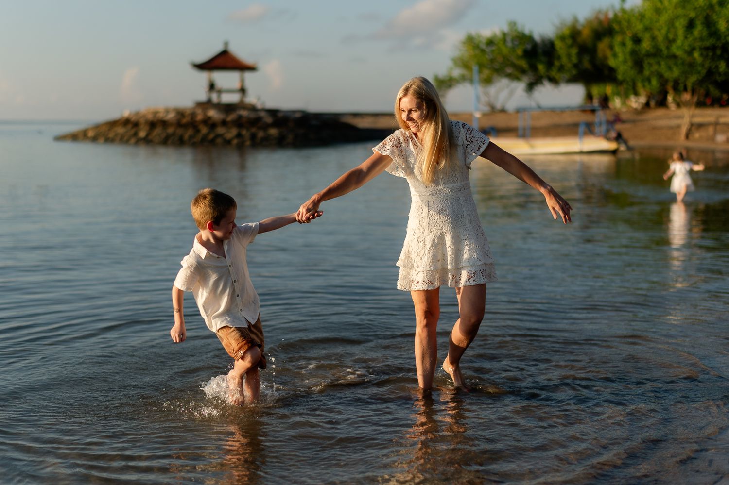 Blonde mother and child splashing through shallow ocean water on a tropical beach at sunset.