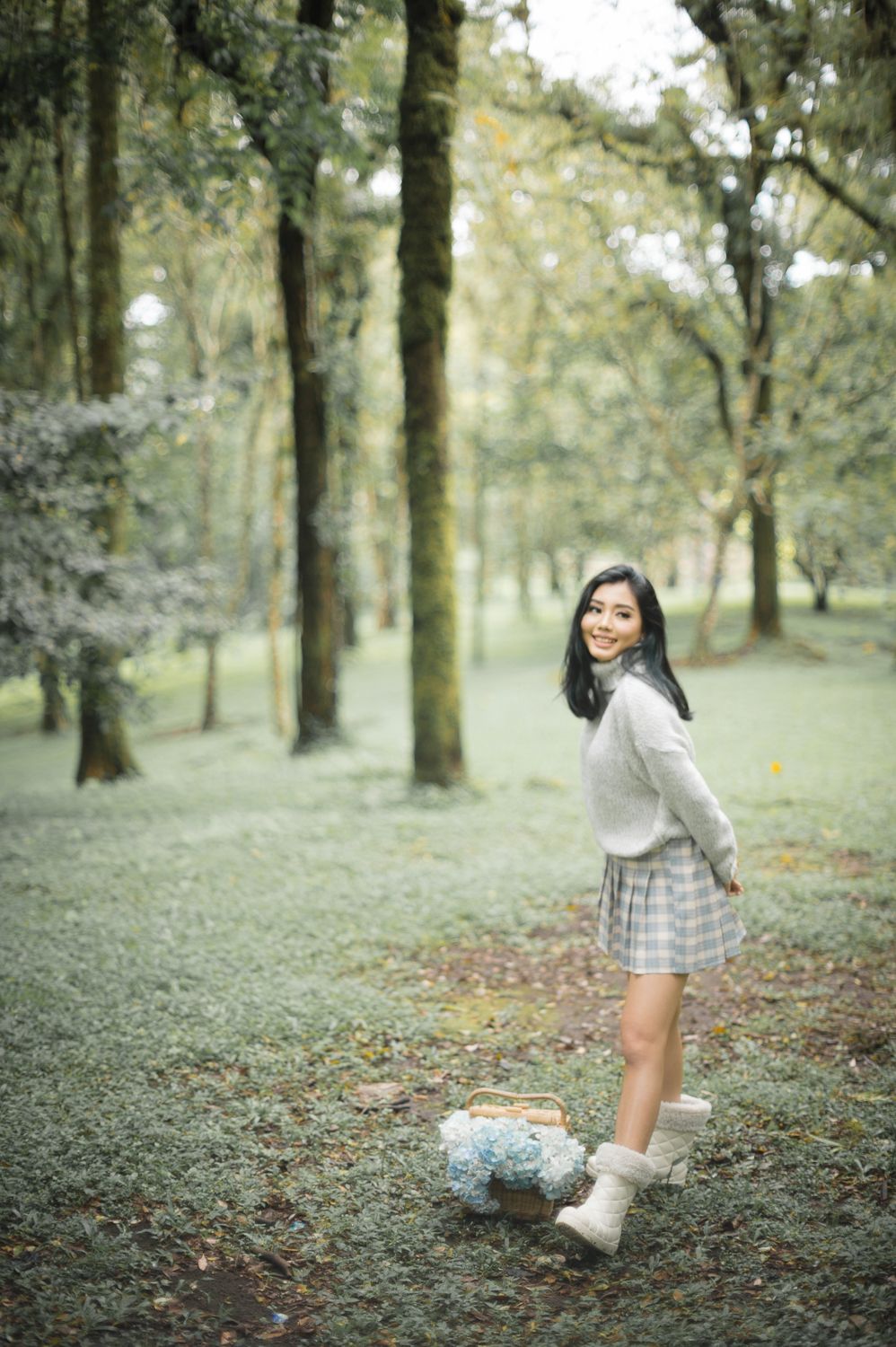 Young Asian woman in plaid skirt and sweater smiling in a lush green forest park holding a flower basket.