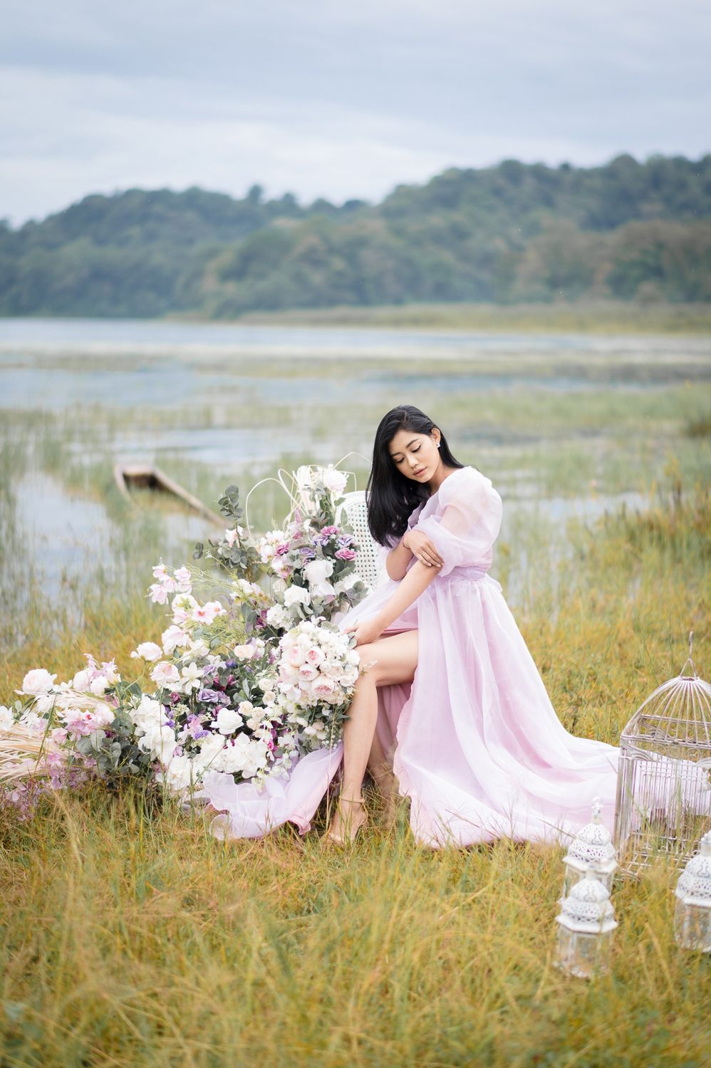 Woman in pink gown sits among flowers by a serene lake, surrounded by lush greenery and a decorative birdcage.