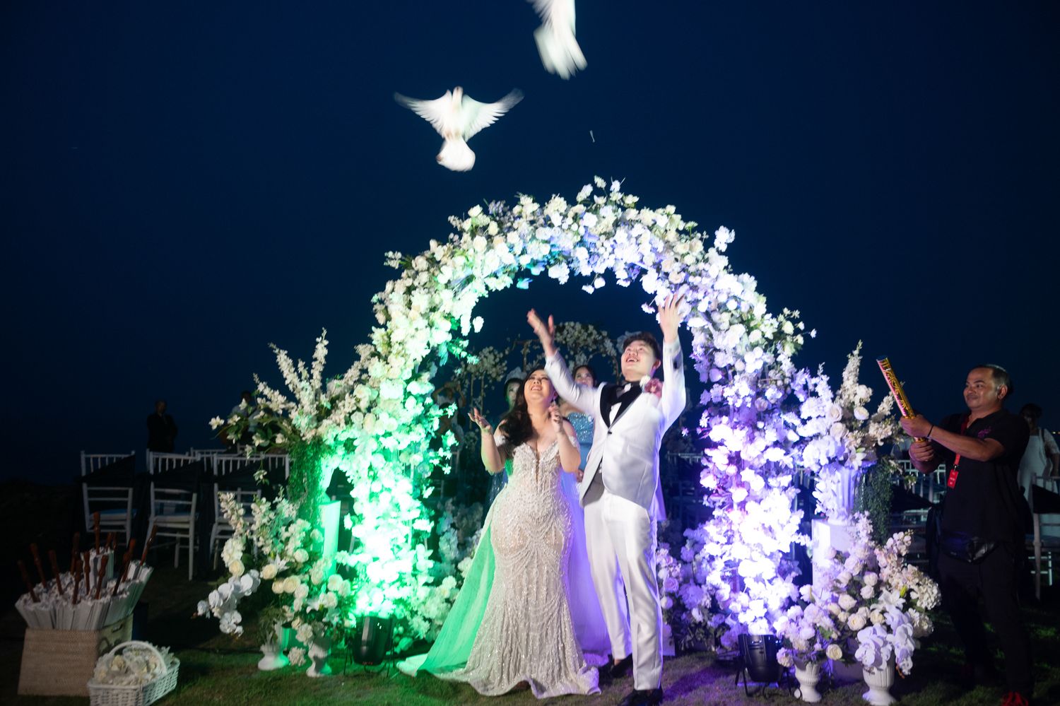 Couple releasing white doves under floral arch at outdoor night wedding ceremony with glowing lights.