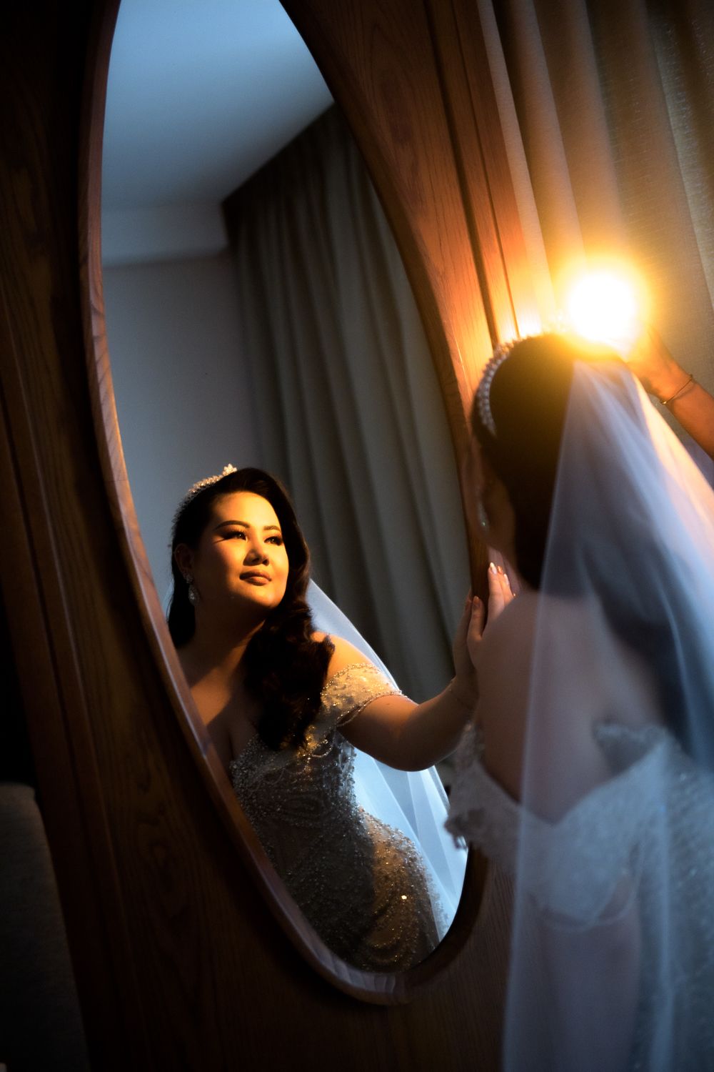 Asian bride in beaded gown admires her reflection in an oval mirror, with warm light glowing beside her veil.