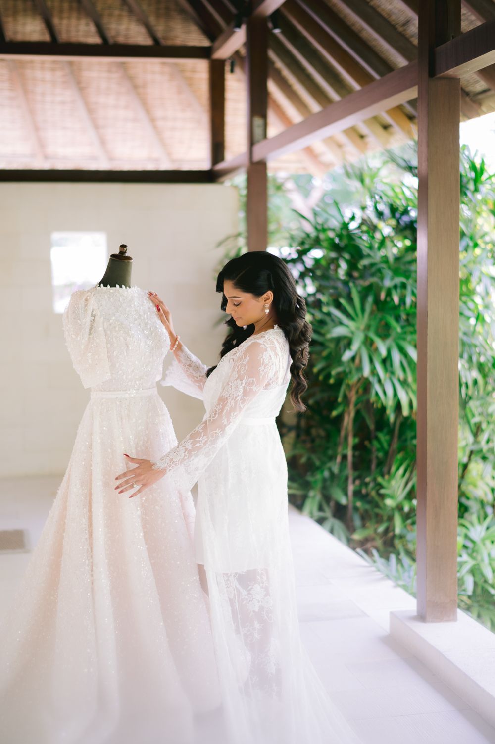 Bride in lace robe adjusts elegant white wedding gown on mannequin in tropical Bali villa setting.