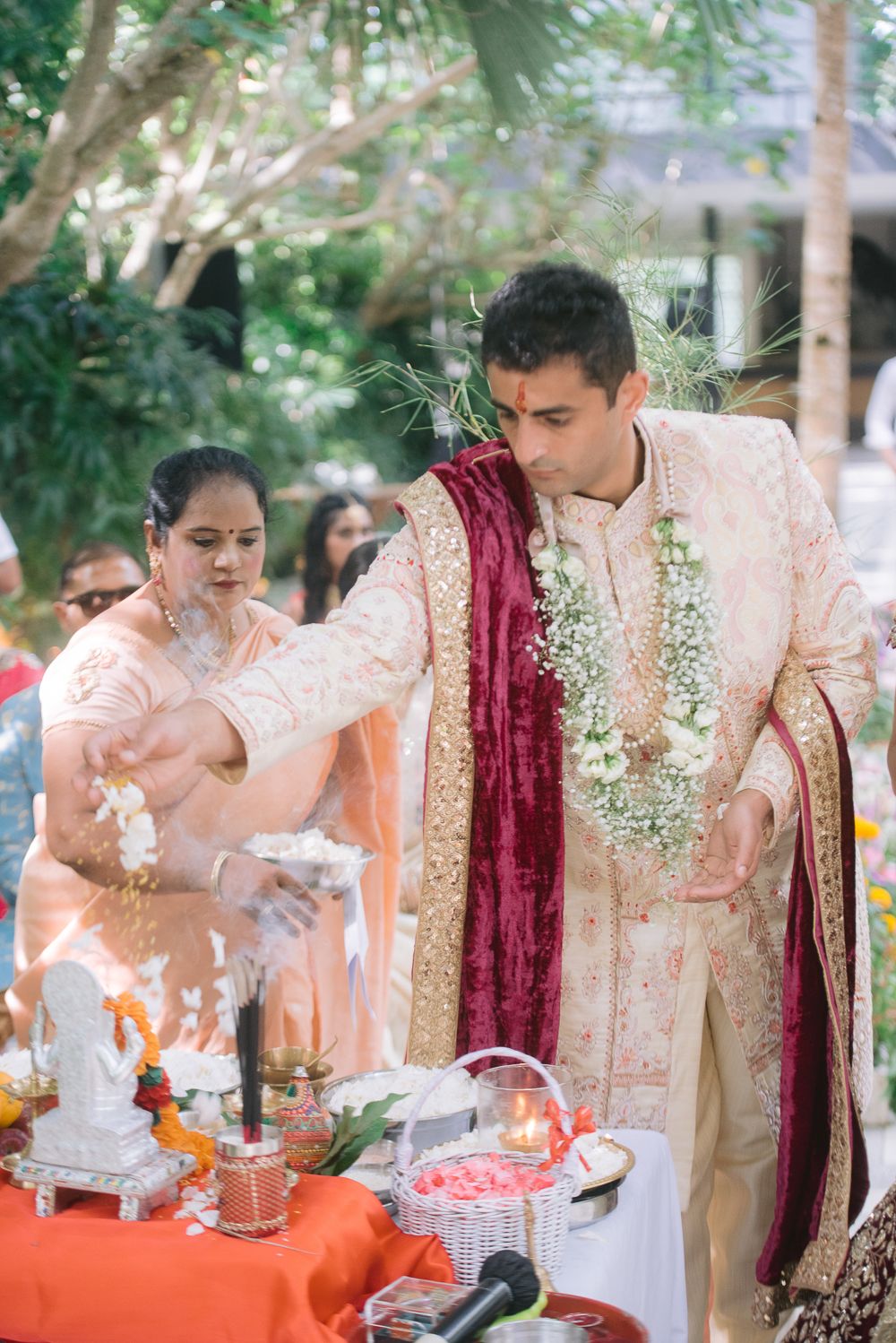 Groom in cream sherwani and red dupatta performs Hindu wedding ritual at outdoor ceremony altar.
