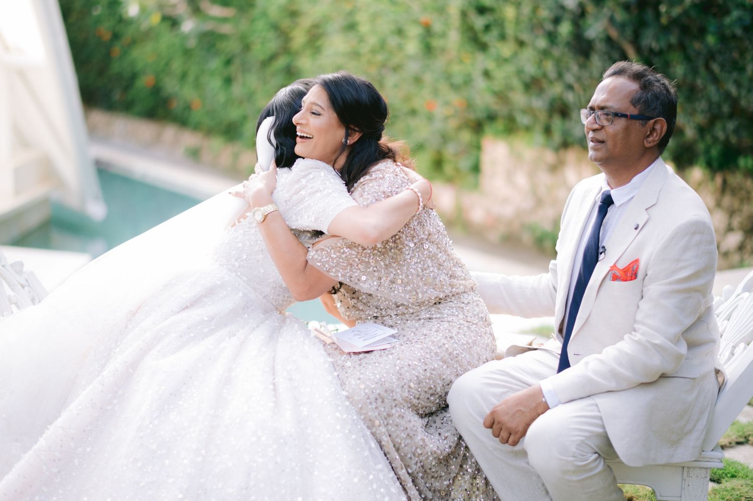 Bride in a beaded gown embracing a woman in a sequined dress while a man in a white suit watches outdoors.