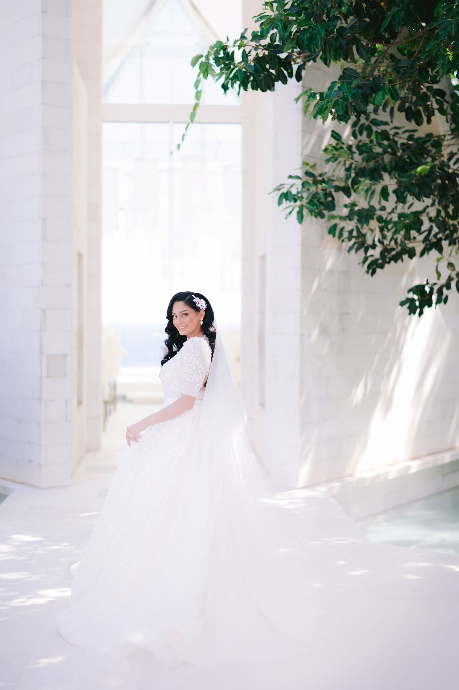 Smiling bride in a flowing white wedding gown stands in a sunlit white hall with green trees and ocean view.