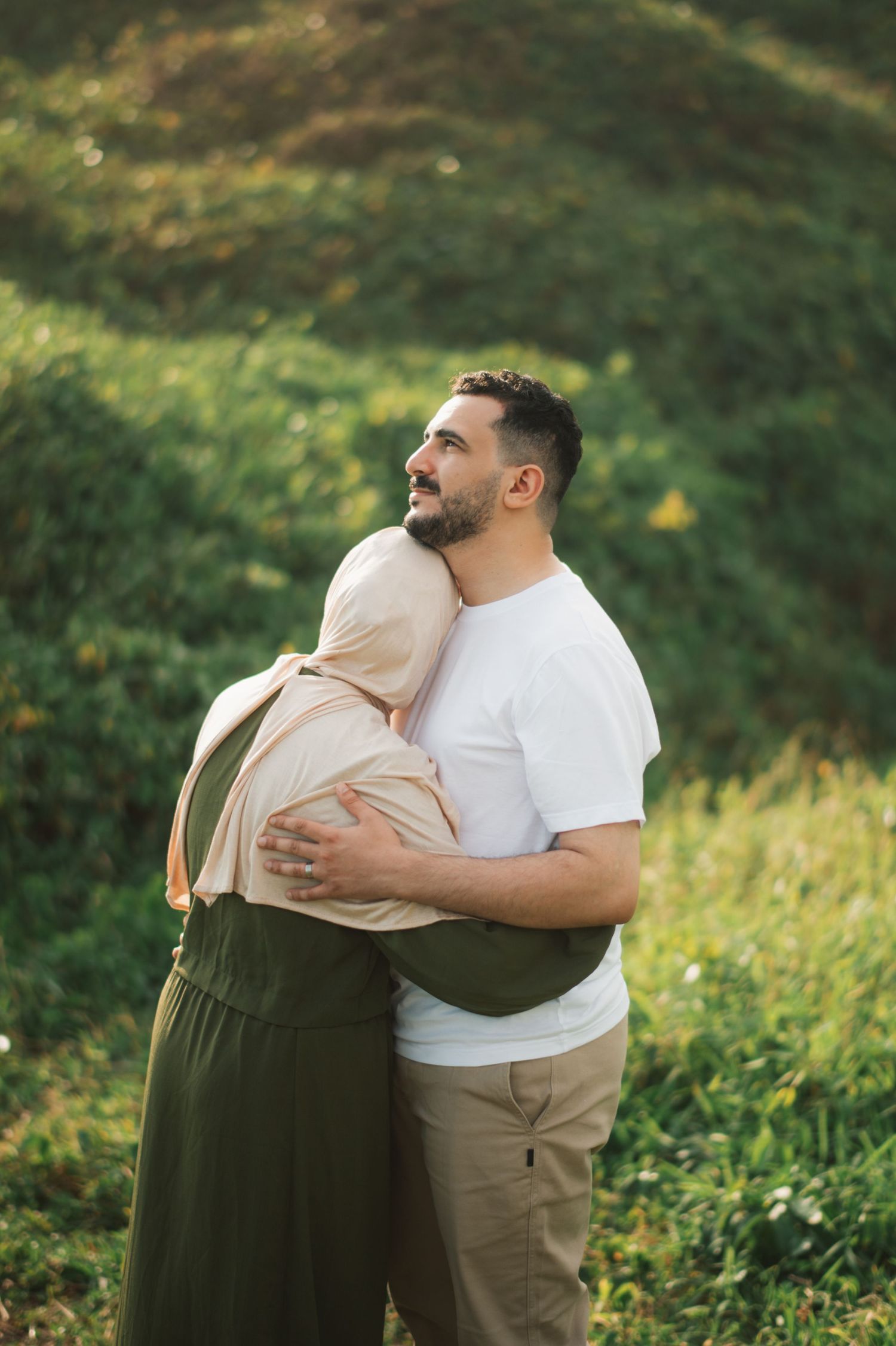 A couple embracing outdoors on a green hillside, woman in hijab and olive dress, man in white t-shirt.