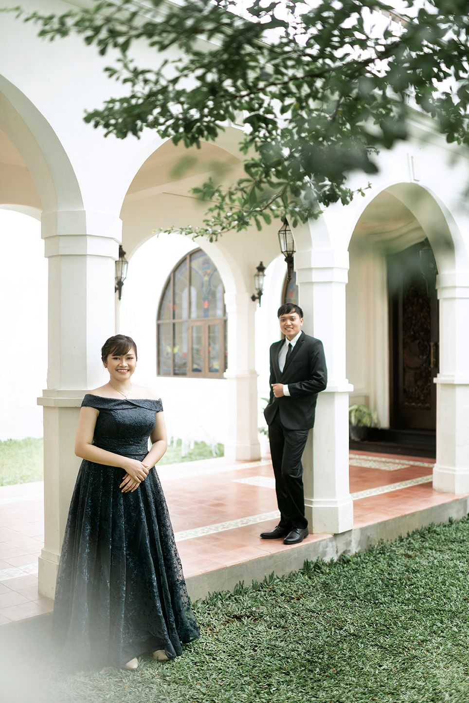 Couple in formal attire posing at elegant white arched building, woman in navy gown, man in black suit.