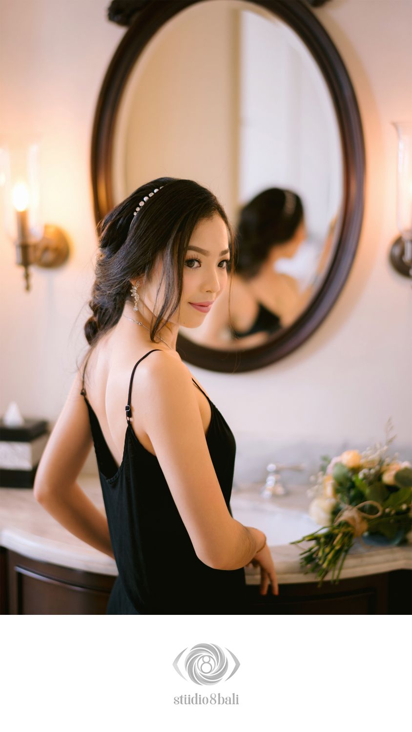 Elegant woman in black dress poses before oval mirror in luxury bathroom with floral bouquet.