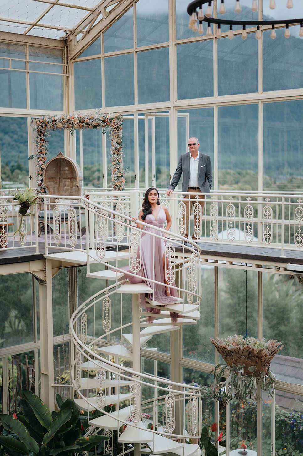 Couple poses on elegant spiral staircase inside a glass greenhouse with mountain views.