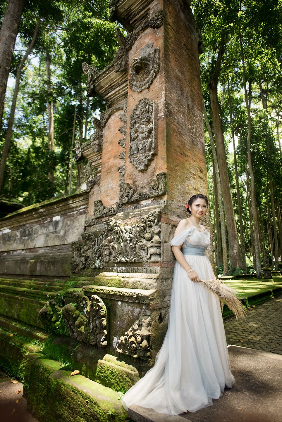 Bride in white gown holds wheat bouquet beside ornate moss-covered Balinese temple wall.