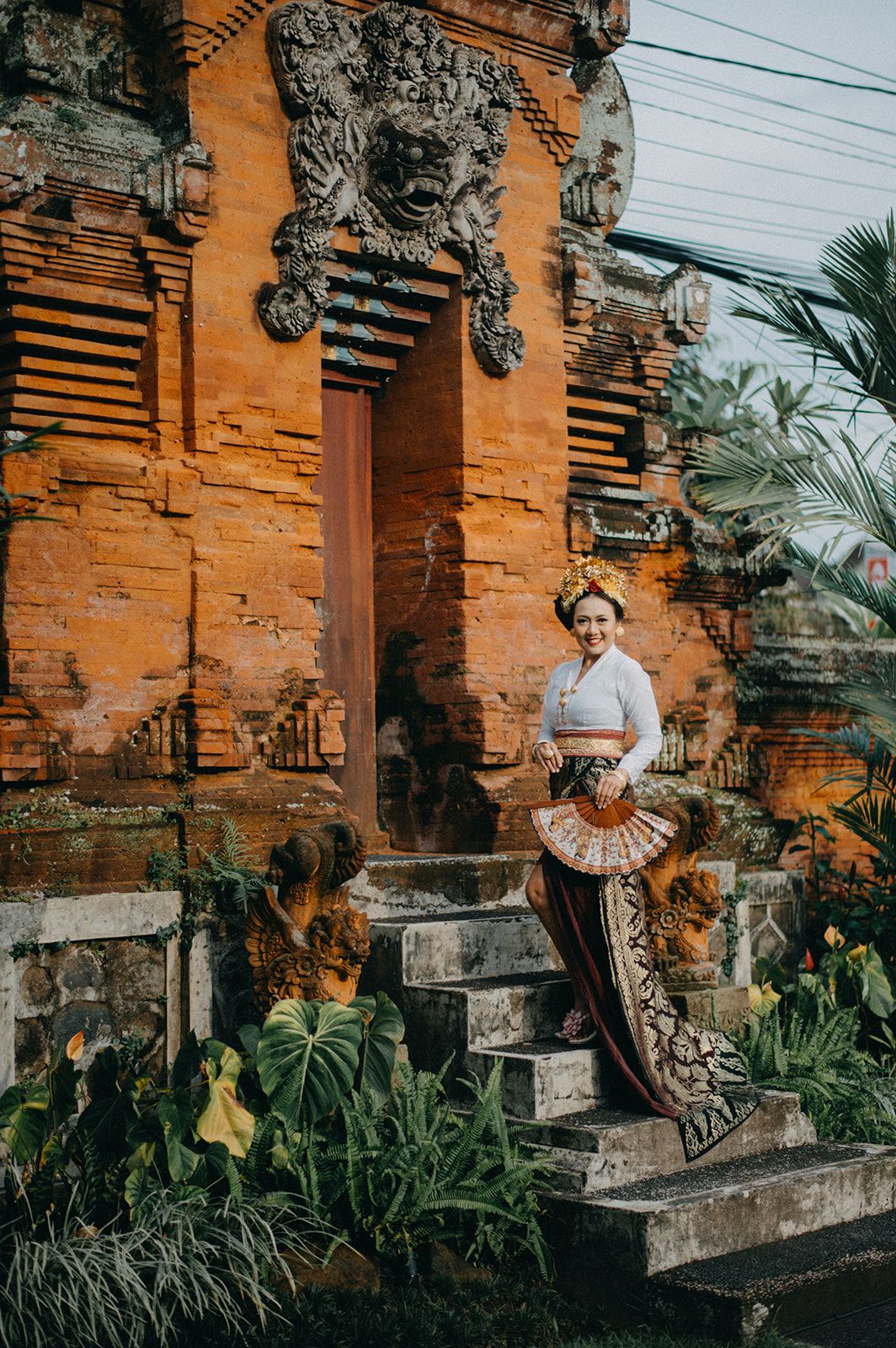 Woman in traditional Balinese attire holding a fan, posing on temple steps with ornate stone carvings.