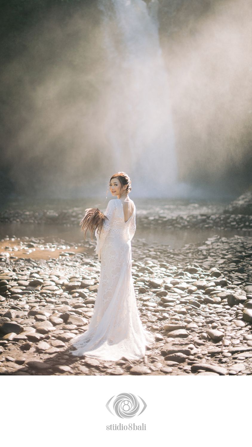 Bride in white lace gown standing on rocky riverbed before misty waterfall in Bali.