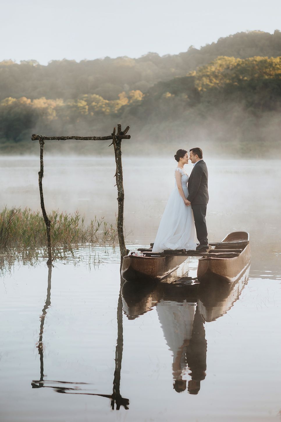 Bride and groom share a romantic kiss on a wooden boat over a misty lake, reflected in calm water.