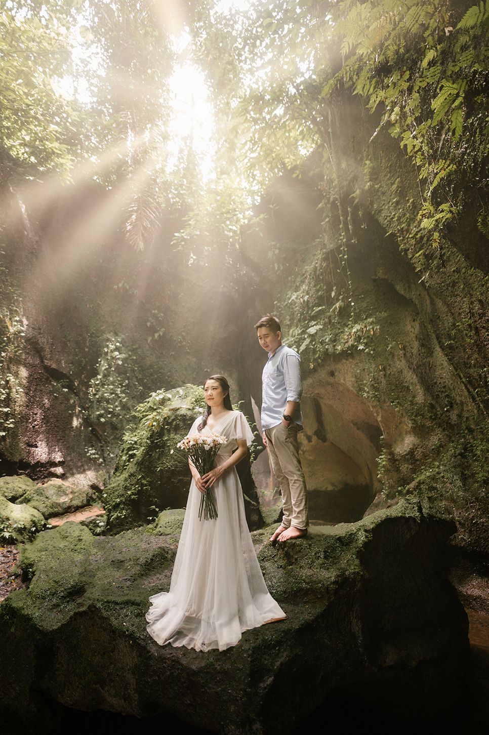 Couple in wedding attire posing on mossy rocks in a lush forest with sunbeams streaming through the canopy.