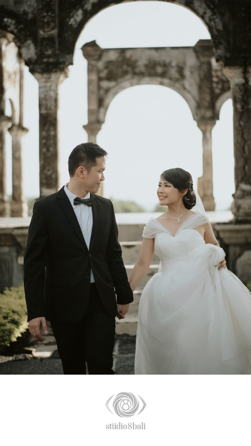 A couple in wedding attire holds hands before stone arches, the groom in a tuxedo and bride in an off-shoulder white gown.