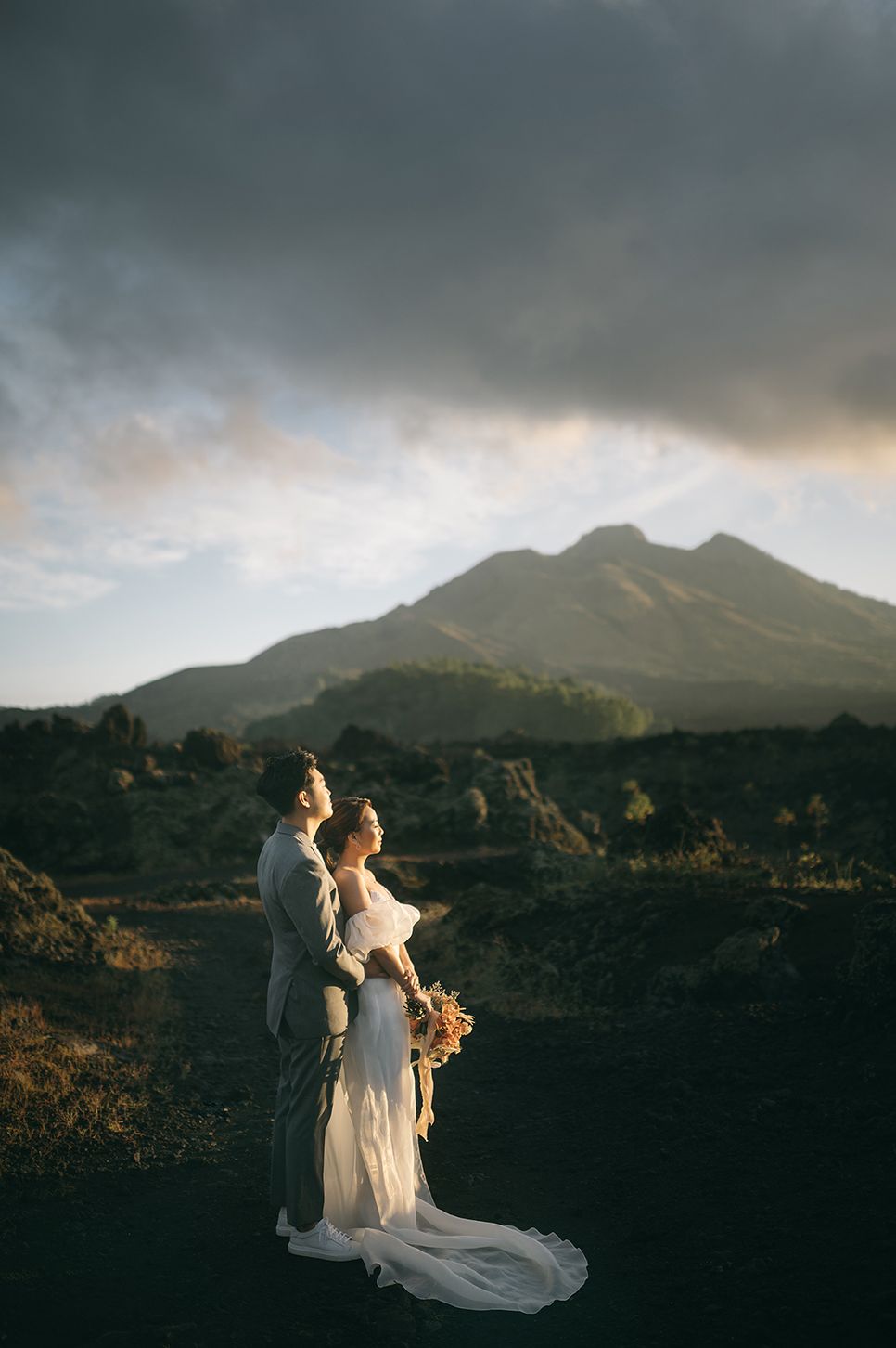 Bride and groom embrace before Mount Batur volcano at sunset in Bali, dramatic clouds overhead.