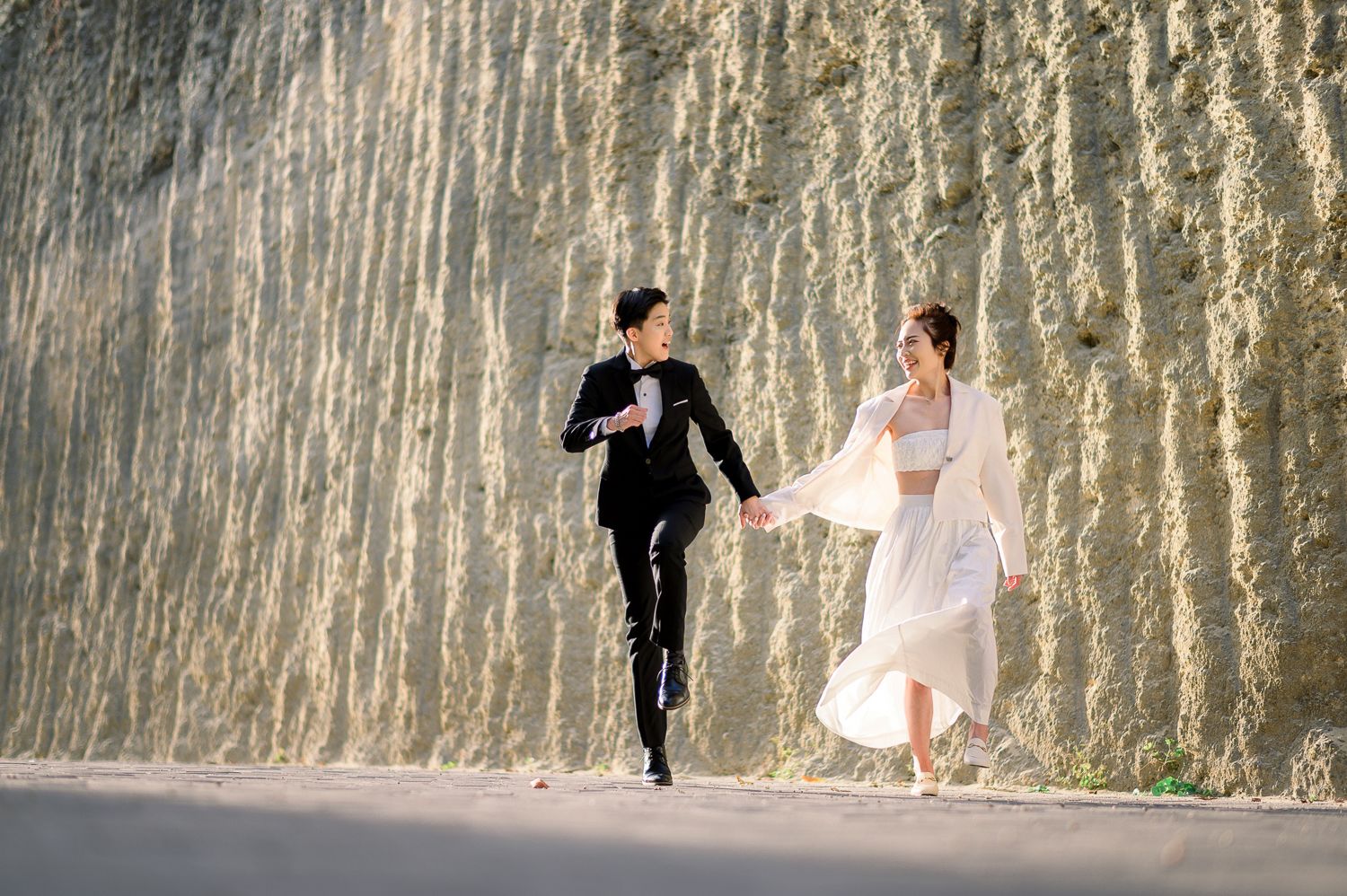 A joyful couple in wedding attire runs hand-in-hand along a sunlit rocky wall backdrop.