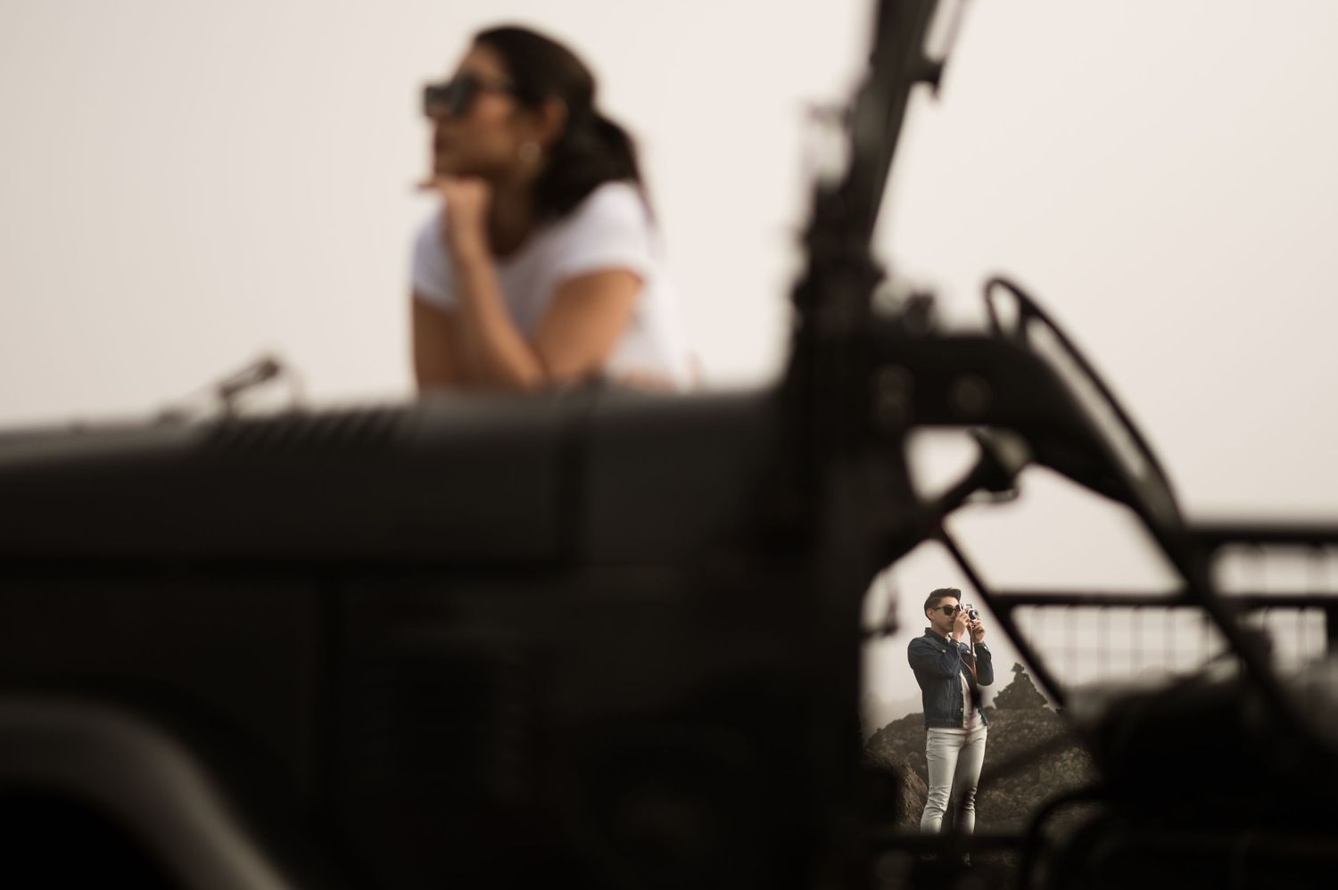 Woman in sunglasses on military vehicle with man photographing in background through gap in equipment.